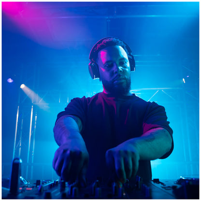 A DJ wearing the AlphaTheta HDJ-F9 Professional Wireless DJ Headphones and a dark t-shirt adjusts a mixing console on a stage under blue and pink stage lighting.