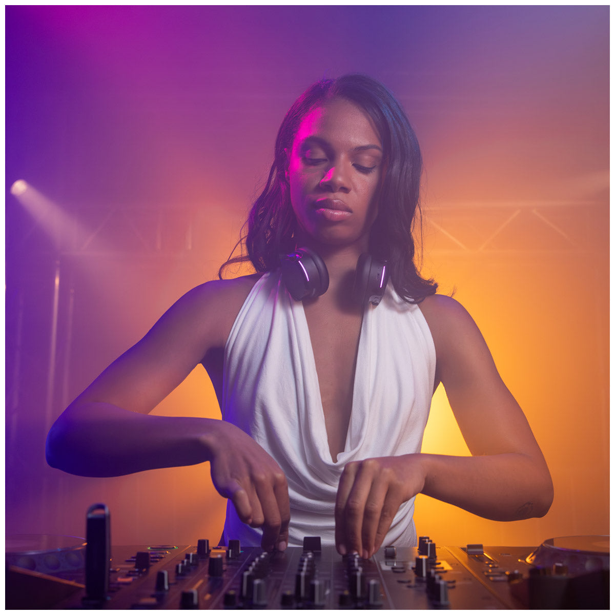 A woman DJing at a mixing console, wearing a white sleeveless top and AlphaTheta HDJ-F9 Professional Wireless DJ Headphones around her neck, under dramatic purple and orange stage lighting.