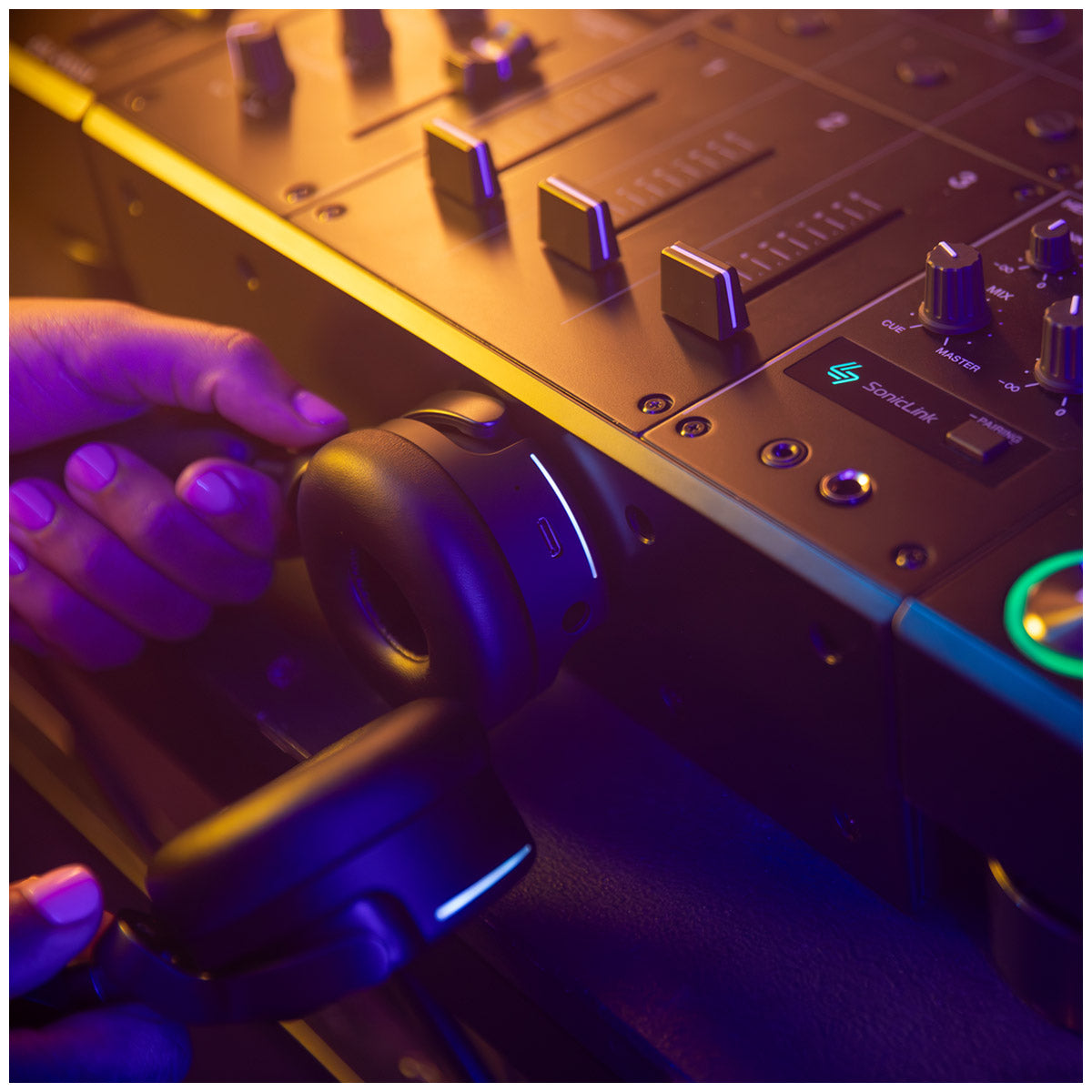 A close-up lifestyle shot of a person's hand holding the AlphaTheta HDJ-F9 Professional Wireless DJ Headphones wirelessly connecting to the DJ mixer with visible controls and knobs under dramatic purple and blue lighting.