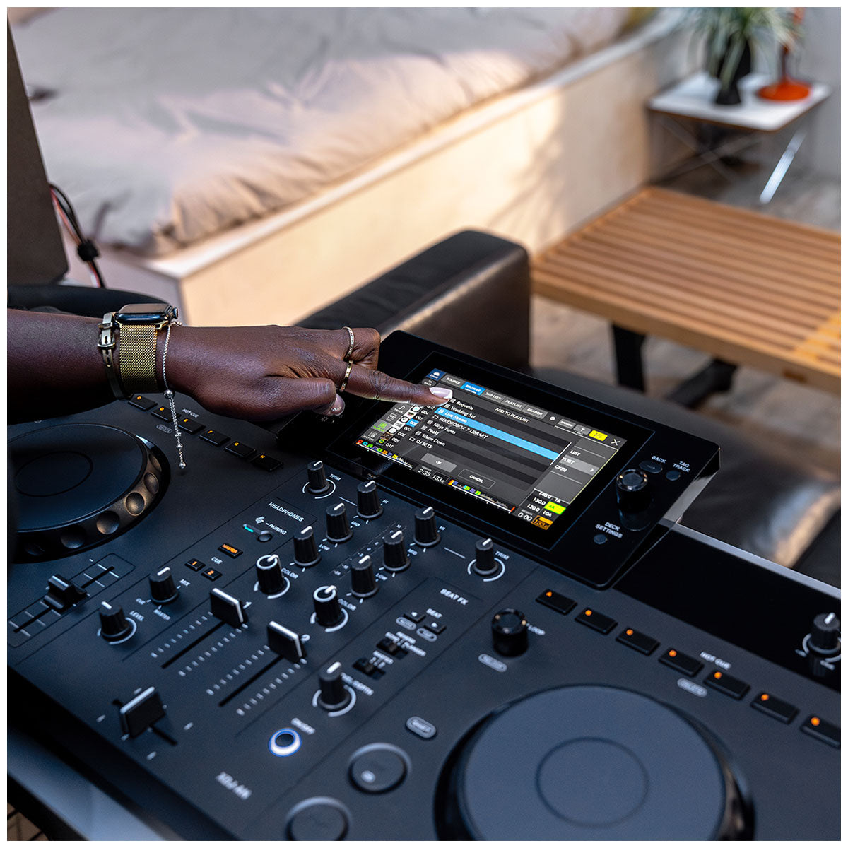 A DJ using a touchscreen controller on a black mixing console in a real indoor setting, with a person's hand and arm visible interacting with the interface.