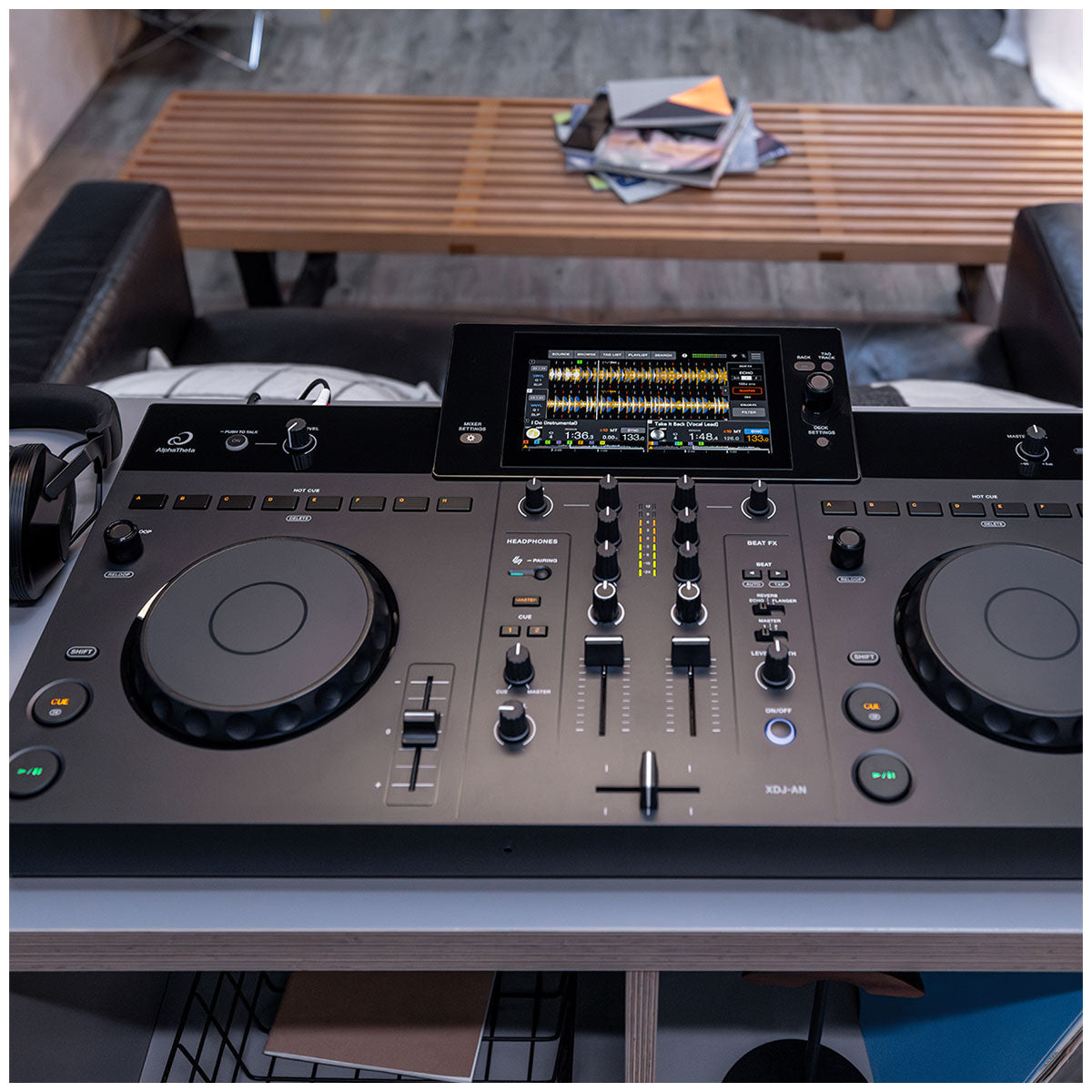 A DJ mixing console with two jog wheels, faders, and a central display screen, photographed in a real indoor environment with furniture and a slatted wooden table visible in the background.