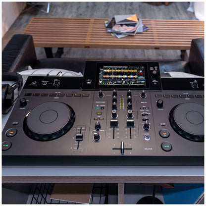A DJ mixing console with two jog wheels, faders, and a central display screen, photographed in a real indoor environment with furniture and a slatted wooden table visible in the background.