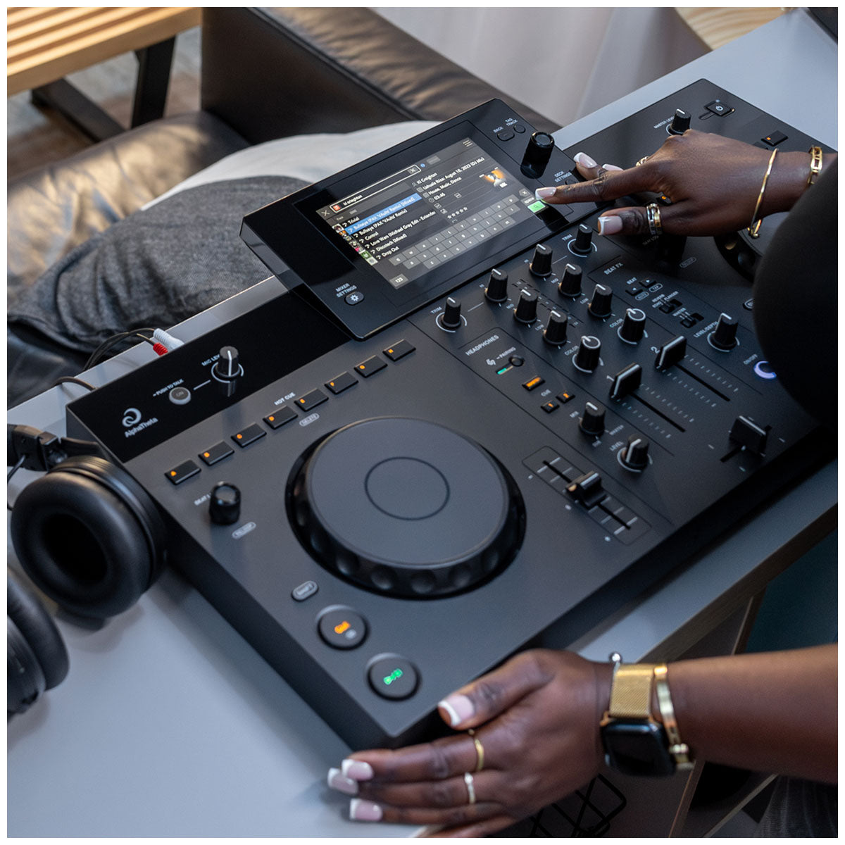 A close-up of a DJ controller mixer with a touchscreen display and knobs, being operated by a person whose hands and arms are visible. Black headphones sit to the left on a gray table in a modern indoor setting.
