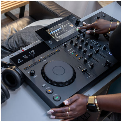 A close-up of a DJ controller mixer with a touchscreen display and knobs, being operated by a person whose hands and arms are visible. Black headphones sit to the left on a gray table in a modern indoor setting.