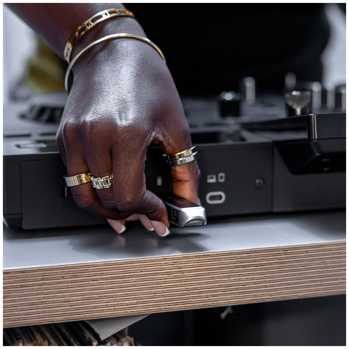 A close-up of a person's hand wearing gold rings and bracelets, holding a small silver USB flash drive in front of a black electronic device on a wooden table.