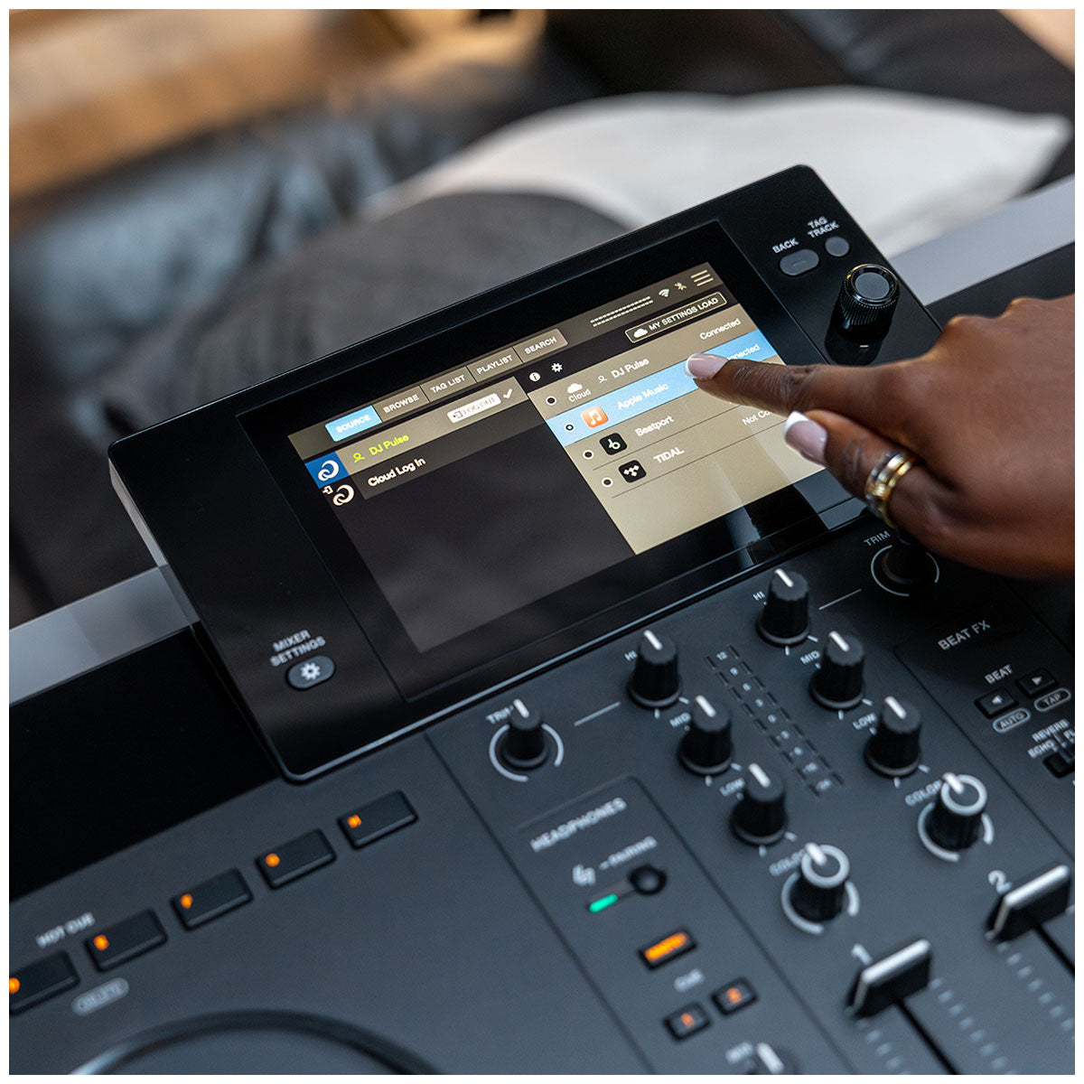 A close-up of a DJ controller with a touchscreen display showing a software interface, with a person's hand touching the screen. The controller has black knobs and sliders, and the background is a blurred indoor setting.